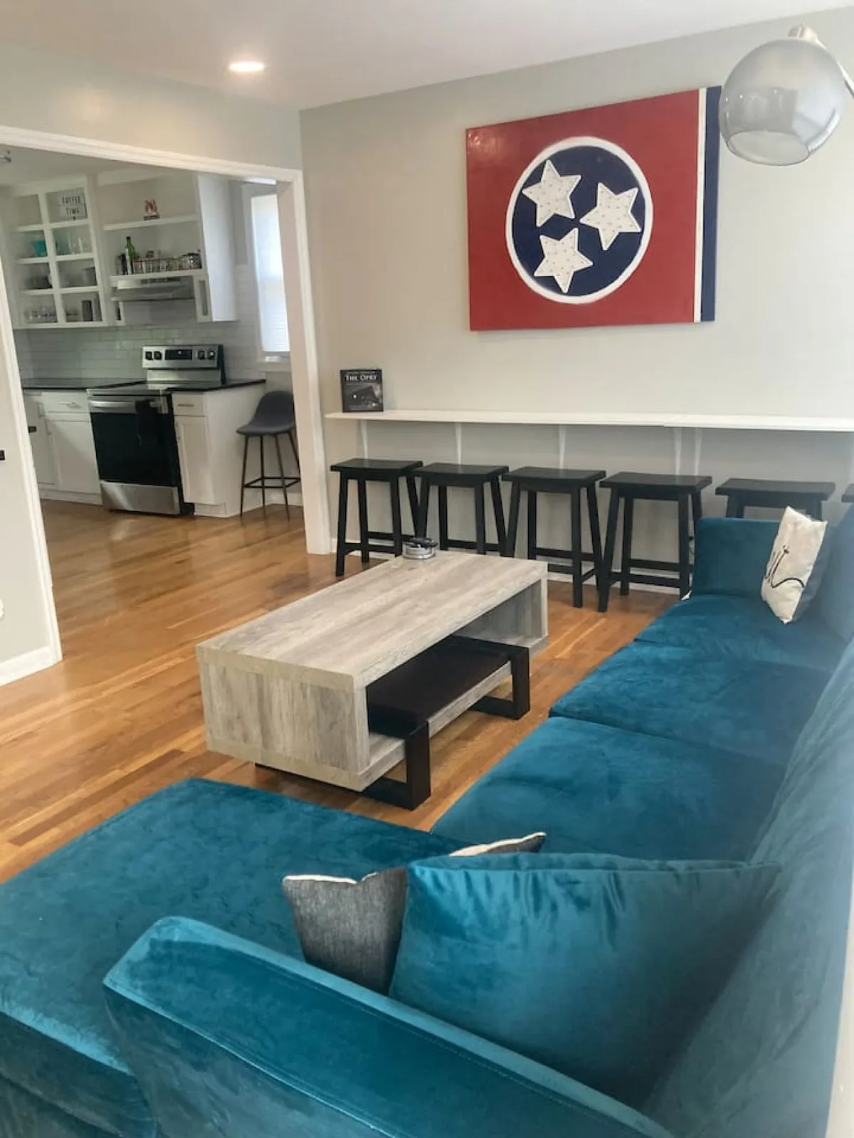 A cozy living area featuring a teal sectional sofa, a wooden coffee table, Tennessee flag artwork on the wall, and bar stools. The space is arranged for socializing and comfortable group gatherings.