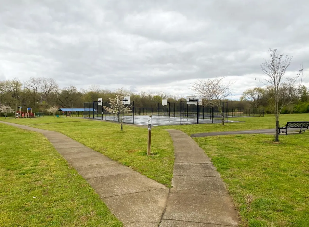 This image captures an outdoor community park area featuring basketball courts, playground equipment, benches, and walking paths. The vibrant activity areas are surrounded by green space, inviting families and children to play and explore.