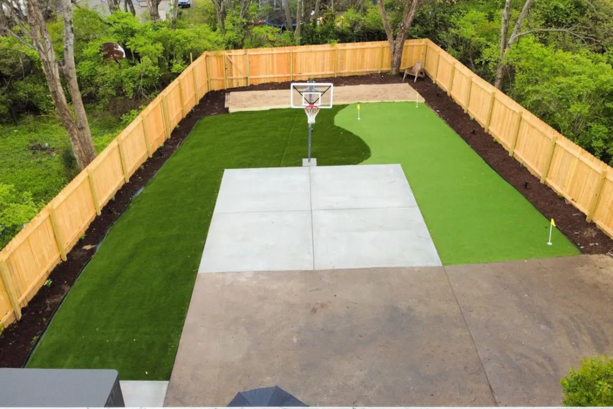 A vibrant outdoor area with a basketball hoop, green artificial turf, and a spacious concrete play area, ideal for energetic basketball games and outdoor gatherings.