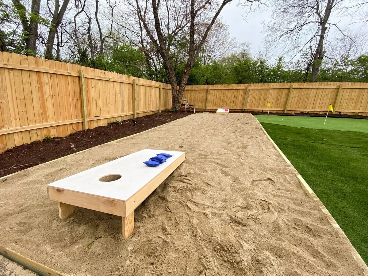 A backyard scene with a sand pit, dedicated cornhole setup, green putting green, and a fenced area, all set up for social and recreational activities. People can be seen enjoying cornhole and mini-golf.