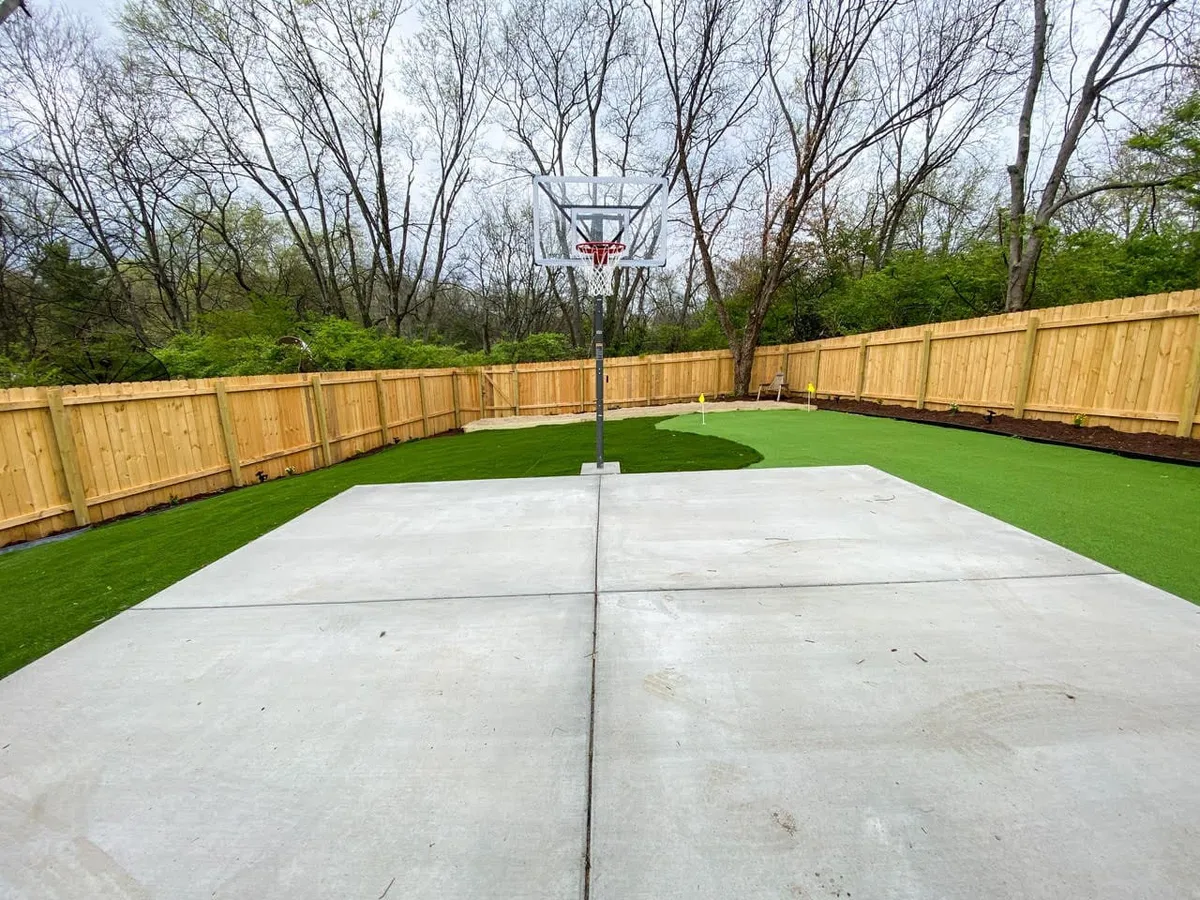 A vibrant exterior shot shows a basketball hoop installed above a concrete court, bordered by lush green artificial grass and enclosed with a wooden fence. The space is inviting, with plenty of room for outdoor activities and team games.