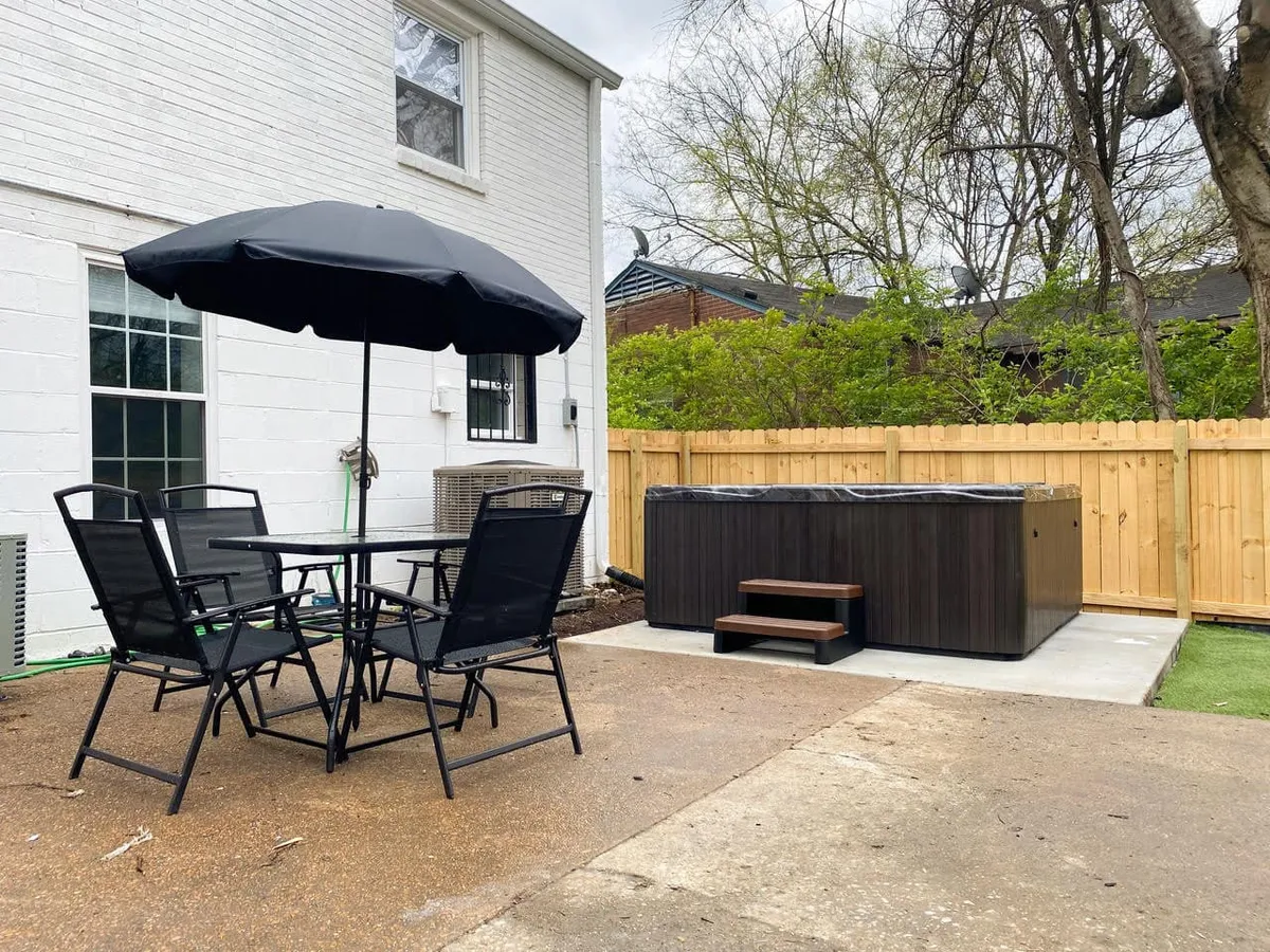 Featuring a large black patio umbrella over a spacious outdoor dining table, with a hot tub and a backyard fence in view. The area exudes comfort, offering ample seating and a relaxing ambiance for groups.