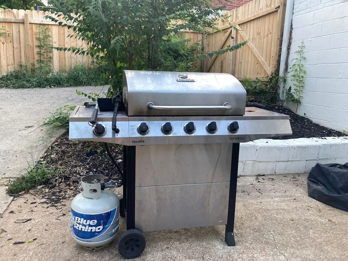 This image centers on a propane grill set amidst lush greenery, with a visible propane tank and an inviting, natural backdrop. The setup suggests a dedicated grilling area within the backyard, tailored for outdoor cooking and socializing.