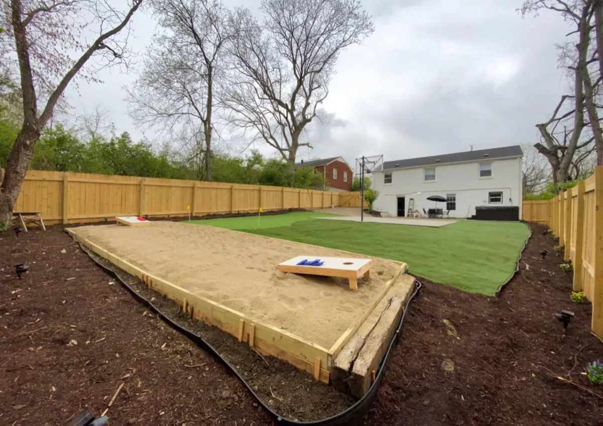 A vibrant backyard scene with a green lawn, sand pit for games like cornhole, and a comfortable outdoor seating area. The setup is open and playful, inviting both adults and children to relax, play, and socialize.