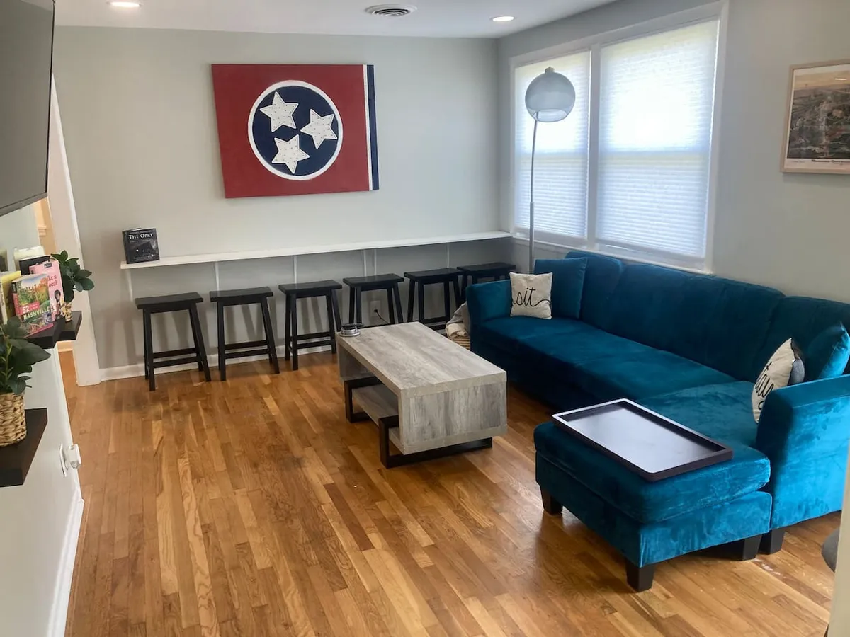 A cozy living room scene with a blue sectional sofa, wooden coffee table, and visible bar stools at a kitchen counter, complemented by Tennessee-themed artwork. The space encourages gathering, relaxation, and lively conversation.