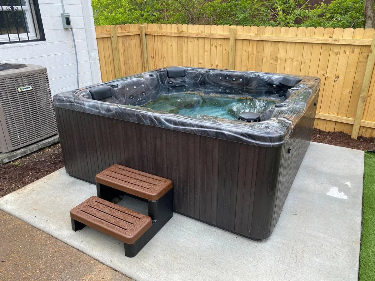 An outdoor hot tub set against a privacy fence with wooden steps leading up. The scene is tranquil and inviting, suggesting a perfect spot for couples to stargaze, friends to unwind, or parents to relax in solitude.